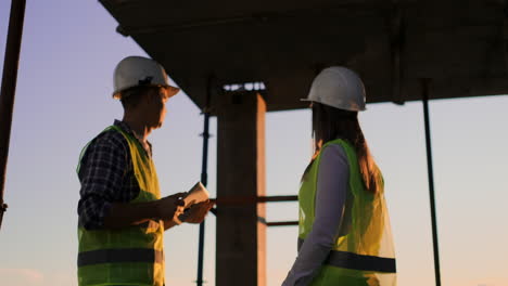 Builder-man-with-a-tablet-and-a-woman-inspector-in-white-helmets-shake-hands-at-sunset-standing-on-the-roof-of-the-building.-Symbol-of-agreement-of-successful-work